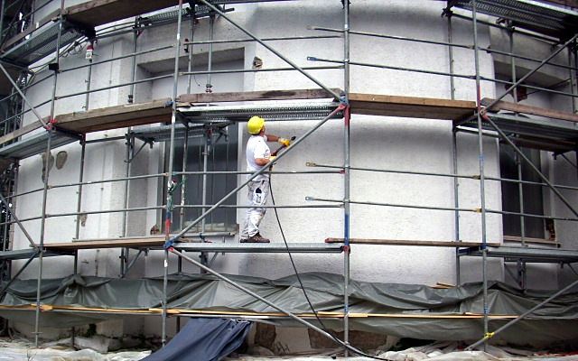 Maler mit Helm beschichtet eine gerundete Hausfassade vom Gerüst aus, unten liegt Abdeckfolie