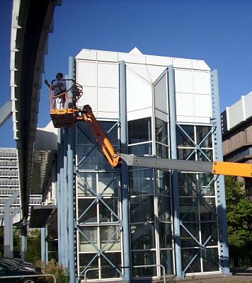 Gläserner Treppenturm, davor der Korb einer orangen Hubarbeitsbühne mit einem Arbeiter