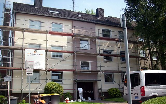 Eingerüstetes Mehrfamilienhaus mit Bauschild, am Eingang arbeitet ein Maler, rechts ein Transporter