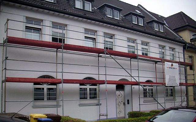 Weißes Wohnhaus mit Rundbogenfenstern und Gauben hinter einem Baugerüst mit Bauschild