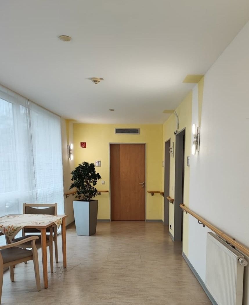 Aufenthaltsbereich am Ende eines Flurs mit Tisch, Stühlen und Pflanze vor gelber Wand