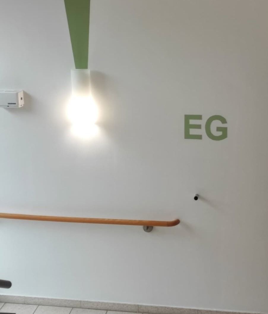 Weiße Wand mit grüner Farbfläche, Wandleuchte, Handlauf und der Beschriftung EG